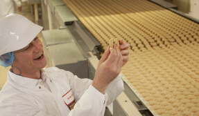 A smiling man wearing a hard hat inspecting a biscuit in a factory